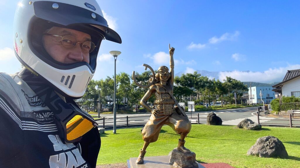 阿蘇駅前のウソップ像の前で撮影された、夏の早朝ツーリング中のライダー。メッシュジャケットとバイザー付きヘルメットを着用し、安全かつ快適な装備で走行。