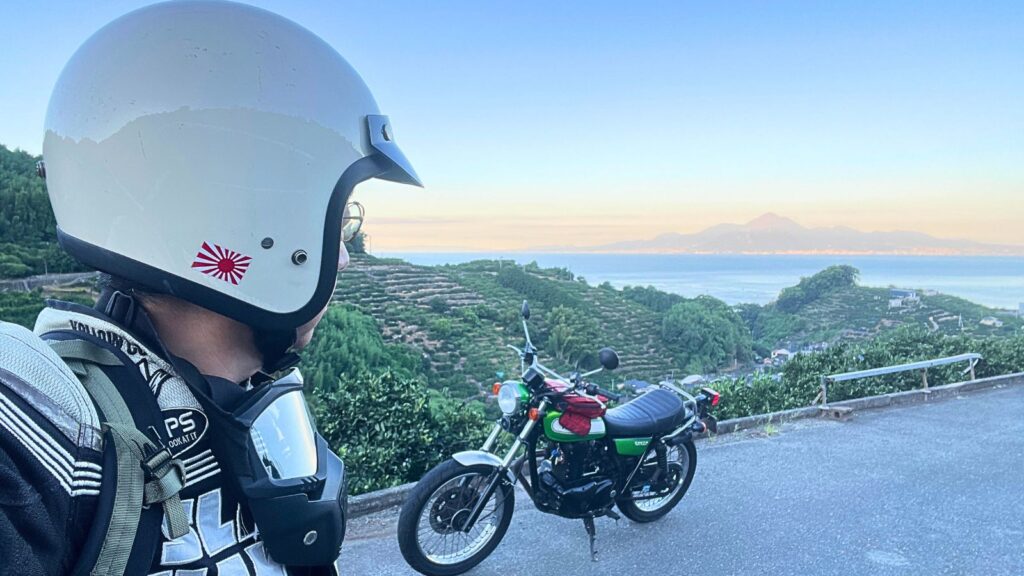 バイク歴40年のライダーが、早朝5時50分に熊本市西区の海沿いで愛機カワサキ250TRと涼しい夏のツーリングを楽しんでいる。背景には有明海越しに朝焼けの雲仙普賢岳が見える。