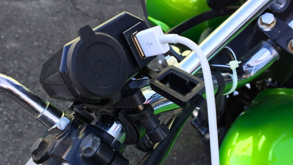 筆者の愛車カワサキ250TRのハンドルに取り付けられたバイク用USB電源。実際にUSBケーブルが差し込まれ、走行中にスマホを充電できる状態を示しており、有線ヘルメットスピーカーシステムのバッテリー問題を解決している。